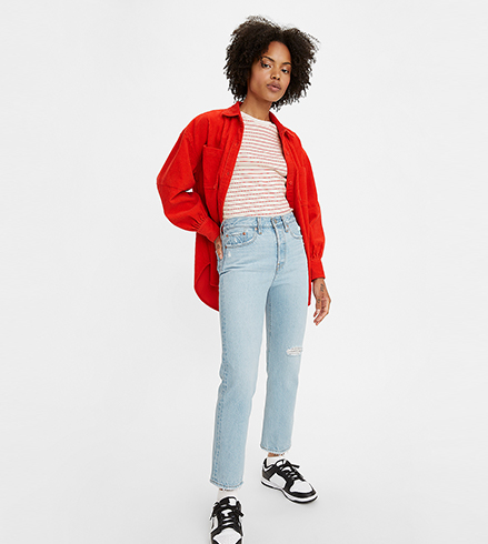 A single model stands in the centre of the image with a dark brown afro hair style wears a red dress shirt over a white and red striped t-shirt which is tucked into jeans. She wears light blue wedgie style jeans that have light distressing at the knee and finishes her looks with black and white sneakers.